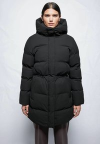 Schwarze Pufferjacke mit hohem Kragen, Knopfverschluss und elastischem Bund. Die Jacke hat ein gesteppter Design und lange, voluminöse Ärmel.