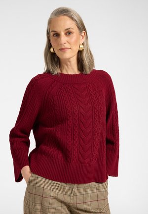 Frau mit grauen Haaren, die goldene Creolen, einen roten Zopfpullover und beige karierte Hosen trägt, steht mit einer Hand in der Tasche.