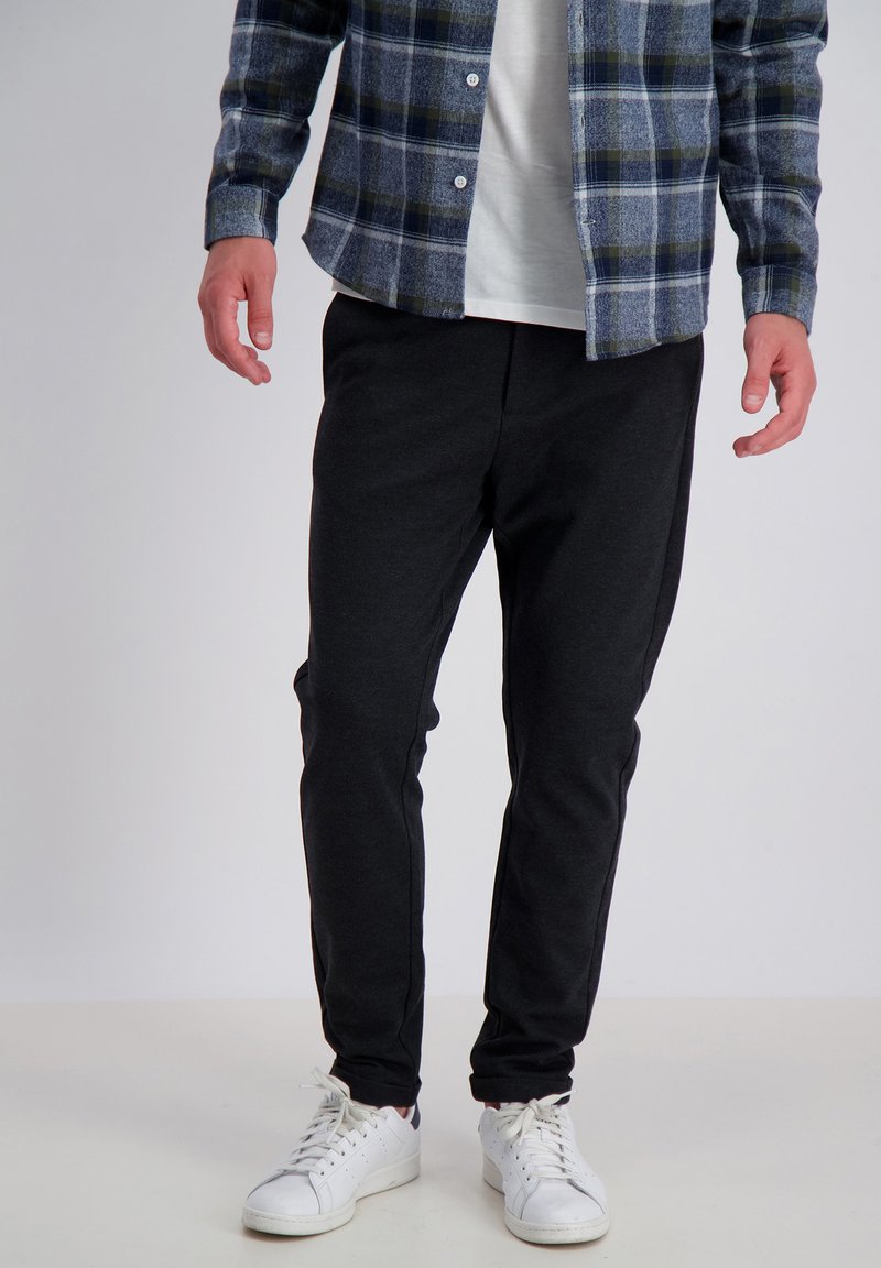 Pantaloni jogger neri con un fit rilassato, realizzati in tessuto morbido. Abbinati a una camicia a quadri blu e sneakers bianche, evidenziando un look casual.