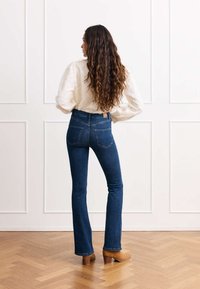 Temno modri flare jeans z visokim pasom, v kombinaciji z strukturirano belo majico. Imajo zanko za pas in subtilne zadnje žepi. Ozadje z leseno talno oblogo in belimi stenami.