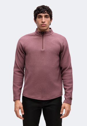 Jonge man met donker krullend haar die een mauve getextureerde trui met lange mouwen en een halve ritskraag draagt, en zwarte broek staat tegen een lichte achtergrond.