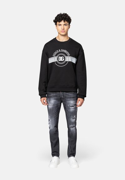Zwarte sweatshirt met een wit logo en strepen, gecombineerd met versleten grijze jeans en witte sneakers. Casual, relaxte pasvorm.