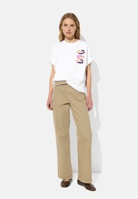 Biała koszulka z krótkim rękawem z kolorowym abstrakcyjnym nadrukiem z przodu, w połączeniu z beżowymi spodniami o szerokich nogawkach i brązowymi butami wsuwanymi.