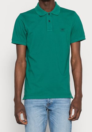 Man draagt een groen poloshirt met korte mouwen en twee knopen, gecombineerd met lichtblauwe spijkerbroek, staand tegen een effen achtergrond.