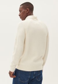 Kreminės spalvos raukšlėta megztinė su aukštu apykakle, ilgomis rankovėmis ir prigludusia apatine dalimi, derinamas su mėlynomis džinsinėmis kelnėmis.