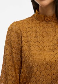 Top in pizzo marrone con un motivo testurizzato caratterizzato da intricati disegni floreali, colletto a volant e maniche lunghe.