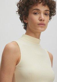 Ribbed, sleeveless turtleneck top in soft cream color, featuring a fitted design with vertical stripes and a high collar.
