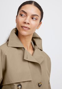 Trench coat beige con ampio colletto, chiusura doppiopetto con bottoni e texture liscia. Cuciture visibili sul colletto.