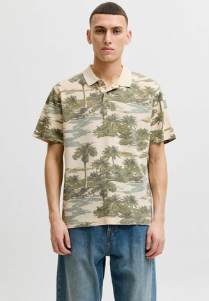 Ung mand iført en beige poloshirt med grøn palmeprint og løse blå jeans, der står mod en ensfarvet lys baggrund.