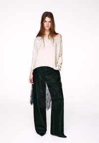 Béžový sveter s odkrytými ramenami, skombinovaný s čiernymi nohavicami s vysokým pásom, ktoré majú priehľadný čipkovaný panel. Textúrovaný, voľného strihu. Čierne špicaté topánky.