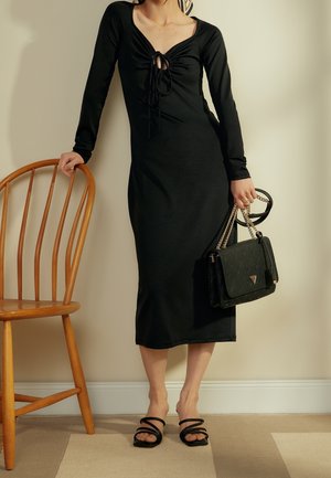 Frau in einem langen schwarzen Kleid mit gerafftem Vorderteil, die eine schwarze Handtasche mit goldenen Ketten hält und neben einem Holzstuhl steht.