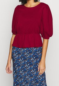 Bordeaux kortærmet top med tekstureret stof og peplum talje, båret over en blomsterprintet blå nederdel med hvide og orange accenter.
