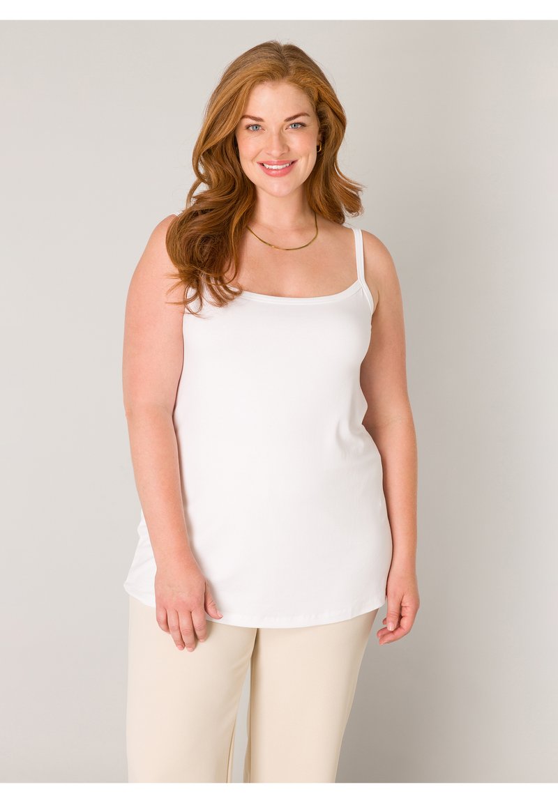 Smiling woman with long red hair wearing a white sleeveless top and beige pants against a plain neutral background.