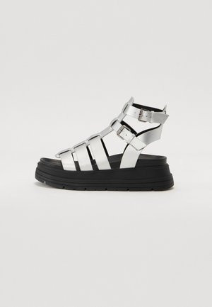 Silberne Faux-Leder-Sandalen mit breiten Riemen und verstellbaren Schnallen, ausgestattet mit einer dicken schwarzen Plateausohle für zusätzlichen Höhe.