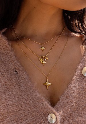 Drie gelaagde gouden kettingen met stervormige hangers, waarvan er één versierd is met kleine bedels, gedragen over een bruine gebreide cardigan.