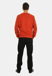 Orange Sweatshirt mit Rundhalsausschnitt und geripptem Saum, kombiniert mit schwarzen Jeans. Der Stoff wirkt weich und hat eine lässige Passform. Keine sichtbaren Muster oder Logos.