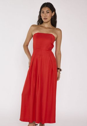 Femme debout portant une robe rouge sans bretelles, longue jusqu’aux chevilles, les mains le long du corps, sur un fond uni clair.