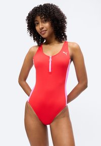 Γυναίκα με σγουρά μαλλιά που φοράει κόκκινο ολόσωμο μαγιό με φερμουάρ μπροστά και λευκό λογότυπο Puma, ποζάροντας με τα χέρια πίσω από την πλάτη.