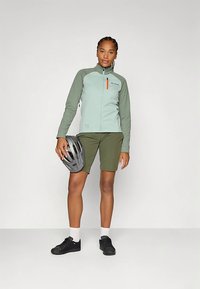 Šviesiai žalia minkšto audinio striukė su užtrauktuku ir pilkais akcentais, derinama su alyvuogių žalių šortų. Juodi dviračių batai ir pilka šalmas rankoje.