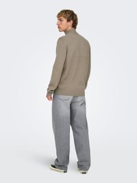 Pull beige à col montant et manches longues, associé à un jean gris clair à jambes larges. Des baskets noires et blanches complètent la tenue.