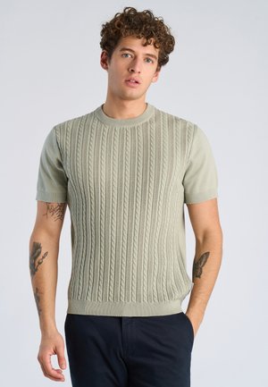 Młody mężczyzna o kręconych włosach, ubrany w jasnoliściasty sweter z krótkim rękawem i ciemne spodnie, pokazujący tatuaże na obu przedramionach.