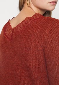 Maglione rosso lavorato a maglia con scollo a V e una delicata rifinitura in pizzo a contrasto sulla schiena, che mette in risalto la texture e i dettagli elaborati.