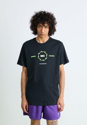 Ung mand med krøllet hår iført en sort Nike T-shirt med grønt logo og lilla shorts, stående foran en ensfarvet lys baggrund.