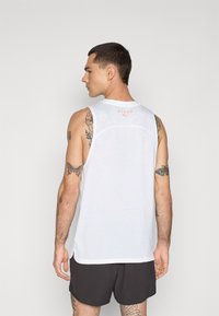 White sleeveless athletic tank top with a mesh texture, featuring a small red logo on the back. Worn with dark shorts, displaying tattoos.