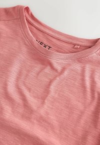 Světle růžové tričko vyrobené z měkké tkaniny, s kulatým výstřihem a jemnou texturou. Na vnitřní straně štítek s nápisem "NEXT".
