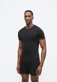 Homme debout devant un fond clair uni, portant un t-shirt noir ajusté et un short noir, face à l'objectif avec une expression neutre.