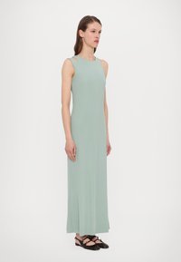 Robe maxi sans manches vert menthe avec un corsage ajusté et des découpes sur les côtés. Le tissu est lisse et fluide, se mariant parfaitement avec des chaussures noires plates.