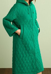 Manteau matelassé vert avec capuche, présentant un motif hexagonal, des boutons-pression et des poches latérales. Confectionné dans un tissu lisse et léger.