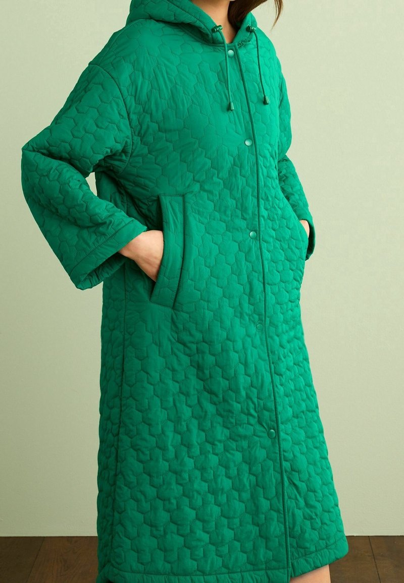 Manteau matelassé vert avec capuche, présentant un motif hexagonal, des boutons-pression et des poches latérales. Confectionné dans un tissu lisse et léger.