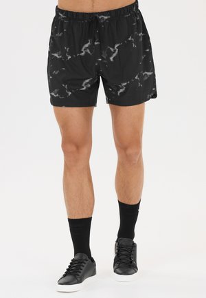 Mandeben iført sorte og grå mønstrede atletikshorts, sorte crew-sokker og sorte sneakers med hvide såler på en ensfarvet baggrund.