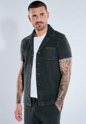 Mann mit Tattoos auf beiden Armen, der ein kurzärmeliges schwarzes gemustertes Hemd über einem weißen T-Shirt trägt und zur Seite blickt.
