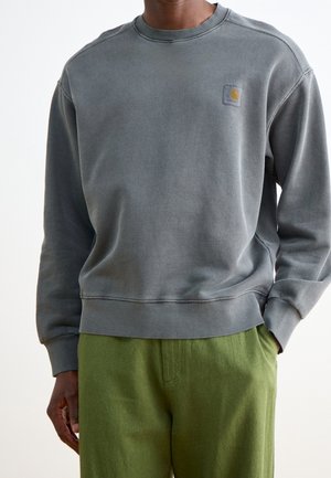 Mann trägt einen ausgewaschenen dunkelgrauen Pullover mit kleinem Logo auf der Brust und olivgrüne Hosen, die Hand in der Tasche, vor schlichtem Hintergrund.