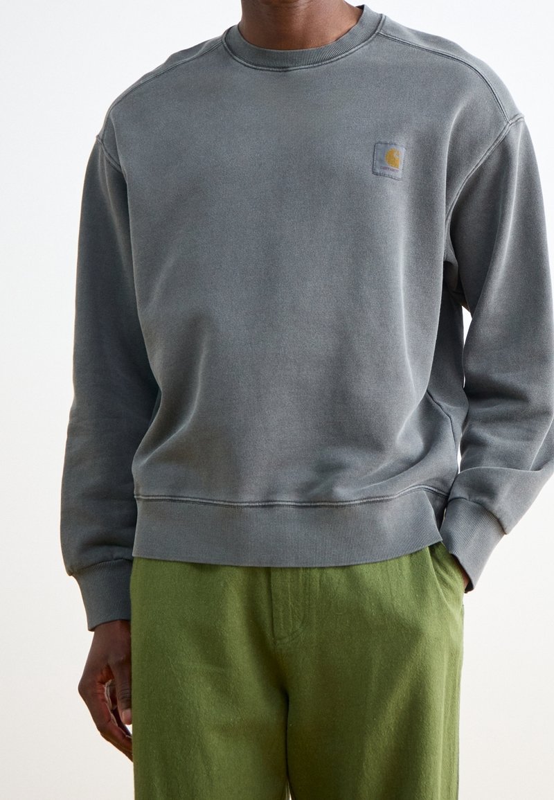 Homme portant un sweat-shirt gris foncé délavé avec un petit logo sur la poitrine et un pantalon vert olive, main dans la poche, sur un fond uni.