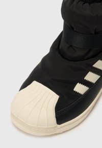 Calzado negro con parte superior acolchada, puño elástico y punta de goma color crema. Presenta tres rayas diagonales en crema texturizado.