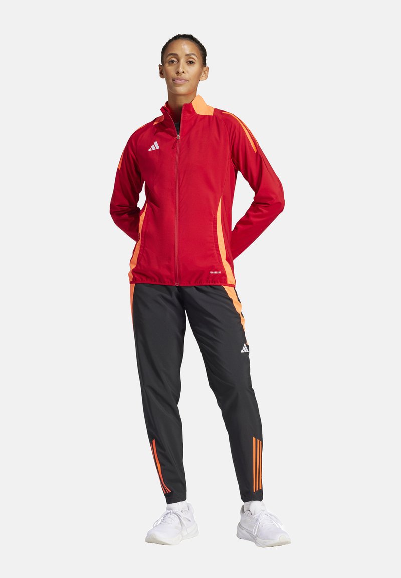 Rote Sportjacke mit orangefarbenen Akzenten, mit Reißverschluss, kombiniert mit schwarzen Hosen mit orangefarbenen Streifen. Hergestellt aus atmungsaktivem Material.