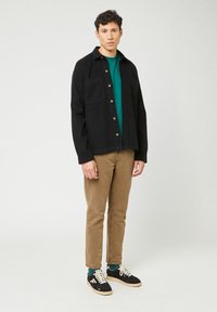 Jonge man die staat, gekleed in een zwart knopenjack over een groene shirt, beige broeken, groene sokken en zwarte sneakers met witte veters.