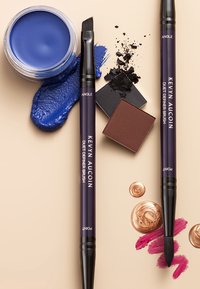Zwei Bürsten mit schräg stehenden schwarzen Borsten, eine blaue Creme in einem Glasgefäß und verschiedene kosmetische Puder in kleinen quadratischen und runden Formen.