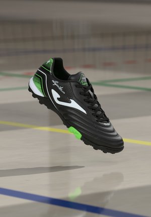 Scarpa sportiva nera con accenti verdi e bianchi, caratterizzata da una tomaia texturizzata e un sistema di allacciatura. La suola presenta modelli di grip prominenti.