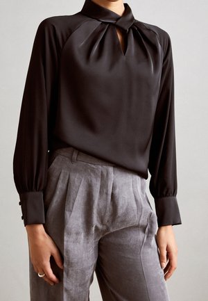 Femme portant un chemisier noir à manches longues avec un décolleté en forme de trou de serrure et un pantalon plissé gris taille haute.