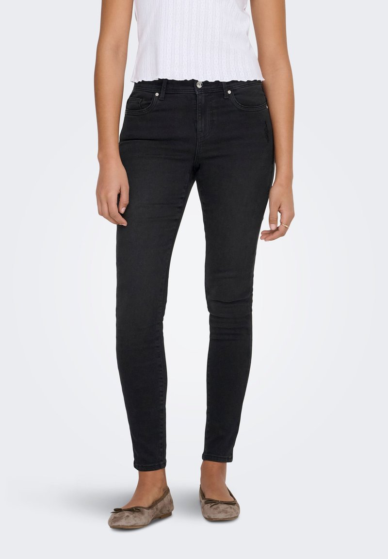 Black denim skinny jeans with five pockets and a mid-rise waistband, paired with a white ribbed top and beige ballet flats.