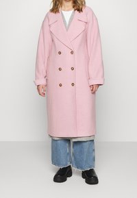 Langer, doppelt geknöpfter rosa Wollmantel mit übergroßen Revers, großen goldenen Knöpfen und Seitentaschen, getragen über einem grauen Pullover und Jeans.