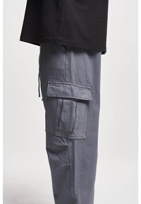 Nahaufnahme von grauen Cargo-Hosen mit Klappentasche am Oberschenkel, getragen mit einem schwarzen Hemd vor einfarbigem Hintergrund.