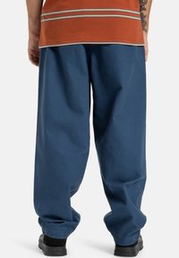 Modré široké kalhoty z měkké látky s uvolněným střihem a manžetami na koncích. Nosené s oranžovým pruhovaným tričkem.