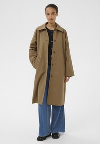 Femme portant un long manteau marron par-dessus un pull noir, un jean bleu à jambes larges et des chaussures noires, debout les mains dans les poches du manteau.