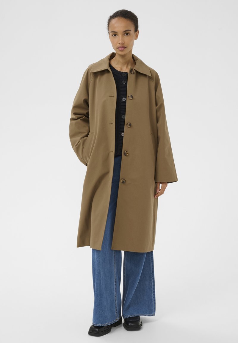 Femme portant un long manteau marron par-dessus un pull noir, un jean bleu à jambes larges et des chaussures noires, debout les mains dans les poches du manteau.