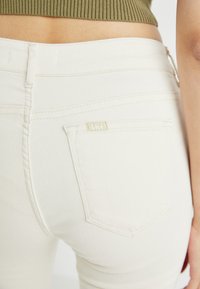 White denim jeans with a smooth texture, featuring a back pocket and subtle stitching. Includes a small logo label above the pocket.